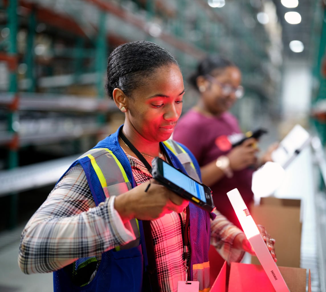 A person wearing a blue safety vest scans an item in a warehouse, while another person in the background also works, holding a clipboard.