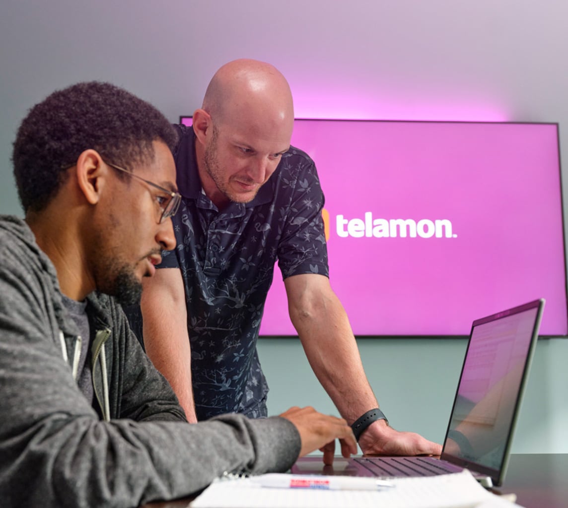 Two men collaborate in front of a laptop. One is sitting and typing, while the other is standing and leans over to look at the screen. A purple Telamon sign is visible in the background.