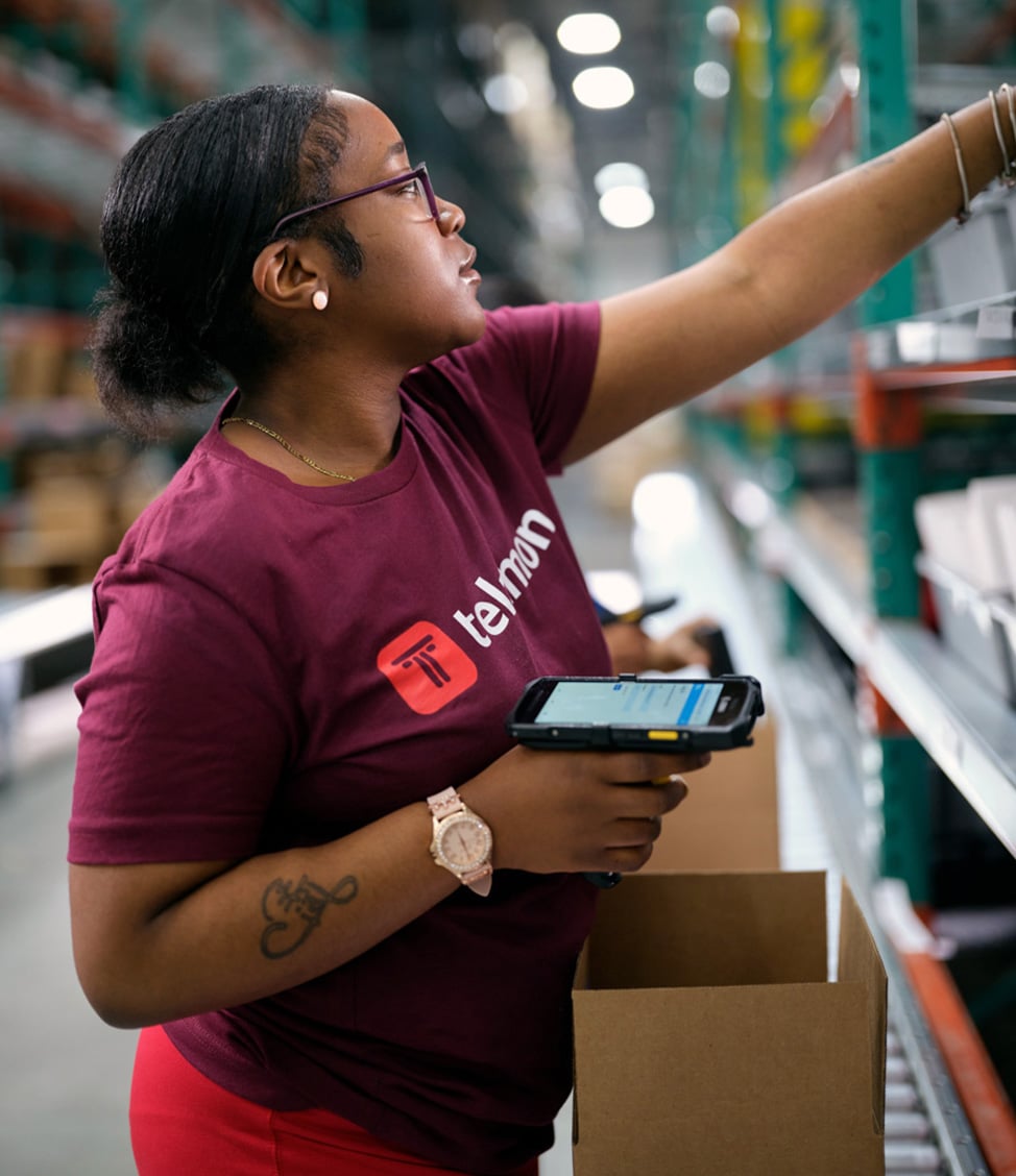 A person wearing a maroon shirt and glasses is using a handheld scanner to pick items from a shelf in a warehouse.