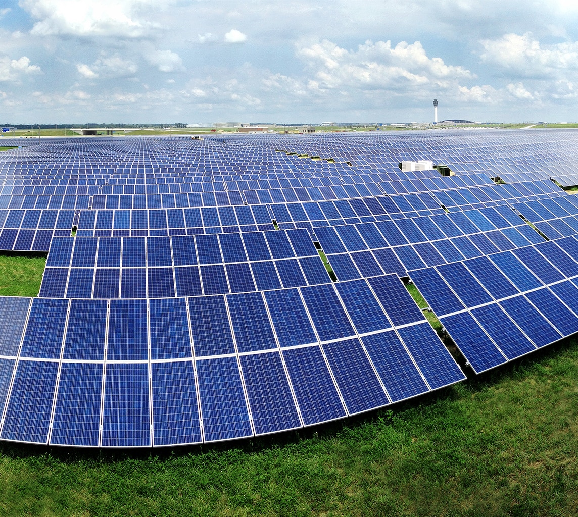 A large solar farm featuring rows of solar panels installed on a grassy area under a partly cloudy sky.