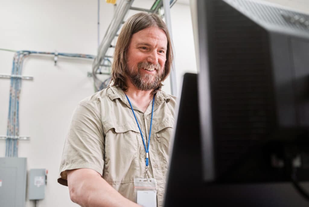 A man with a beard and long hair stands smiling while using a computer in a room with visible electrical wiring. He is wearing a light-colored shirt and a lanyard with an ID badge.