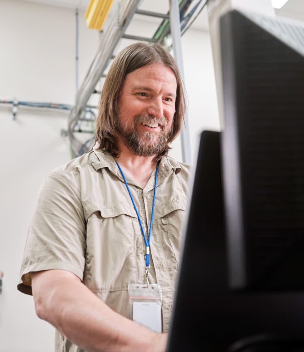 A man with long hair and a beard, wearing a beige shirt and a blue lanyard with an ID, is standing and looking at a computer screen, smiling.