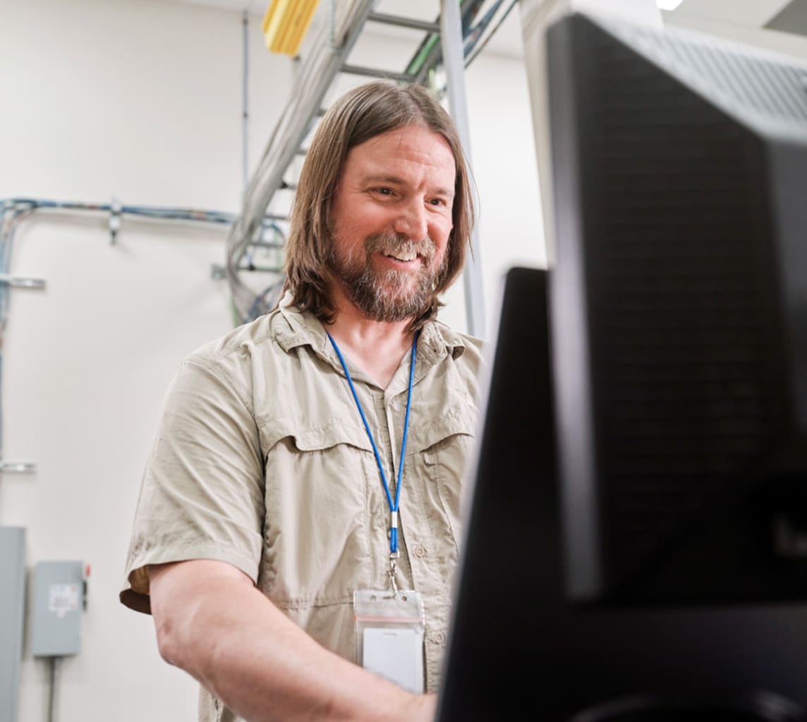 A man with long hair and a beard smiles while working at a computer. He is wearing a khaki shirt and a lanyard with an ID badge. The background shows wires and industrial setup.