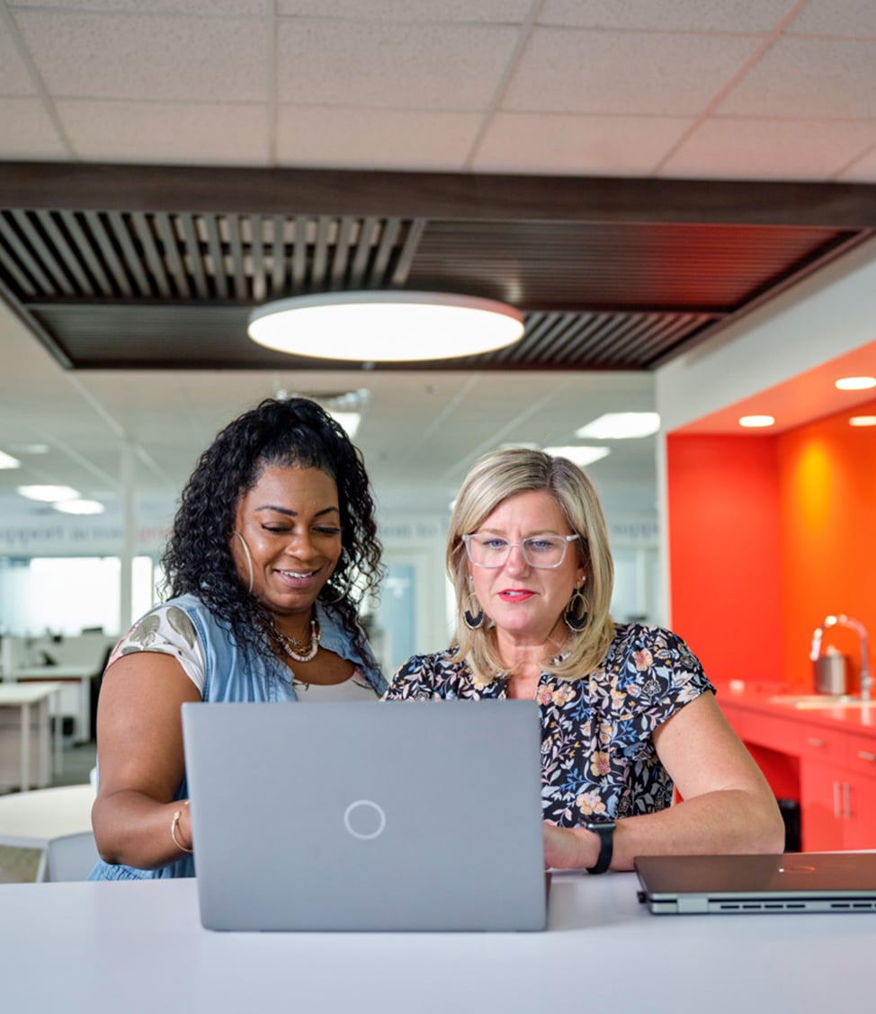 Two women working together at an office desk with a laptop, one standing and the other sitting. The background features a modern office space with bright lighting and an orange accent wall.