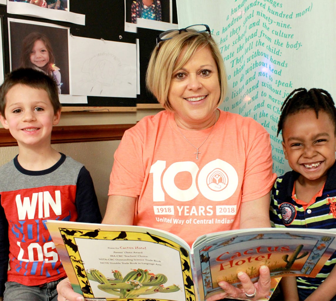 A woman in an orange shirt reading a book to two smiling children, who are sitting on either side of her. A bulletin board with various children's drawings is visible in the background.