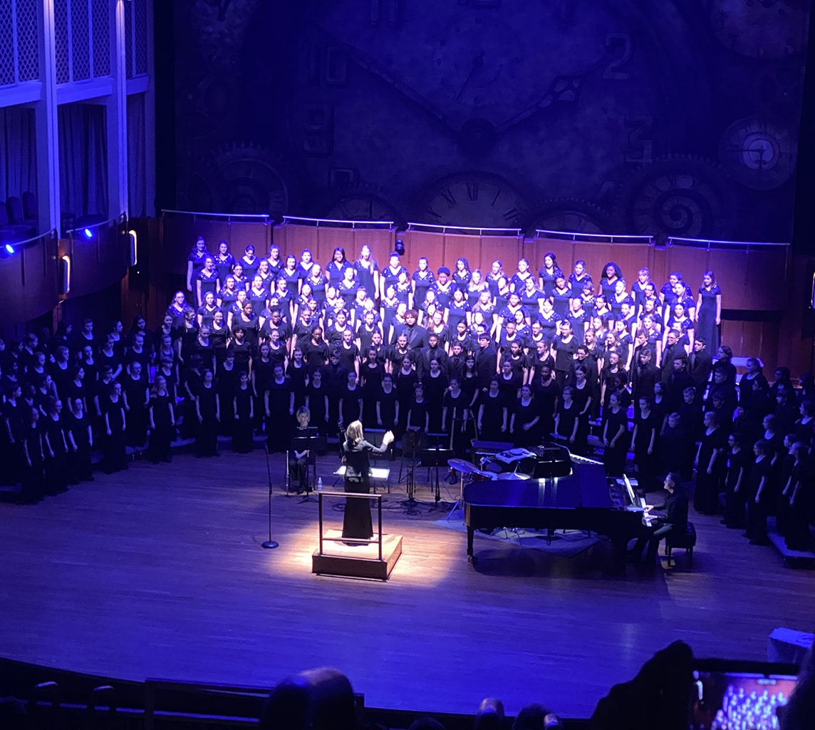 A choir performs on stage with a conductor in front, and a pianist seated at a grand piano. The stage is lit with blue lighting, and a large clock decorates the backdrop.