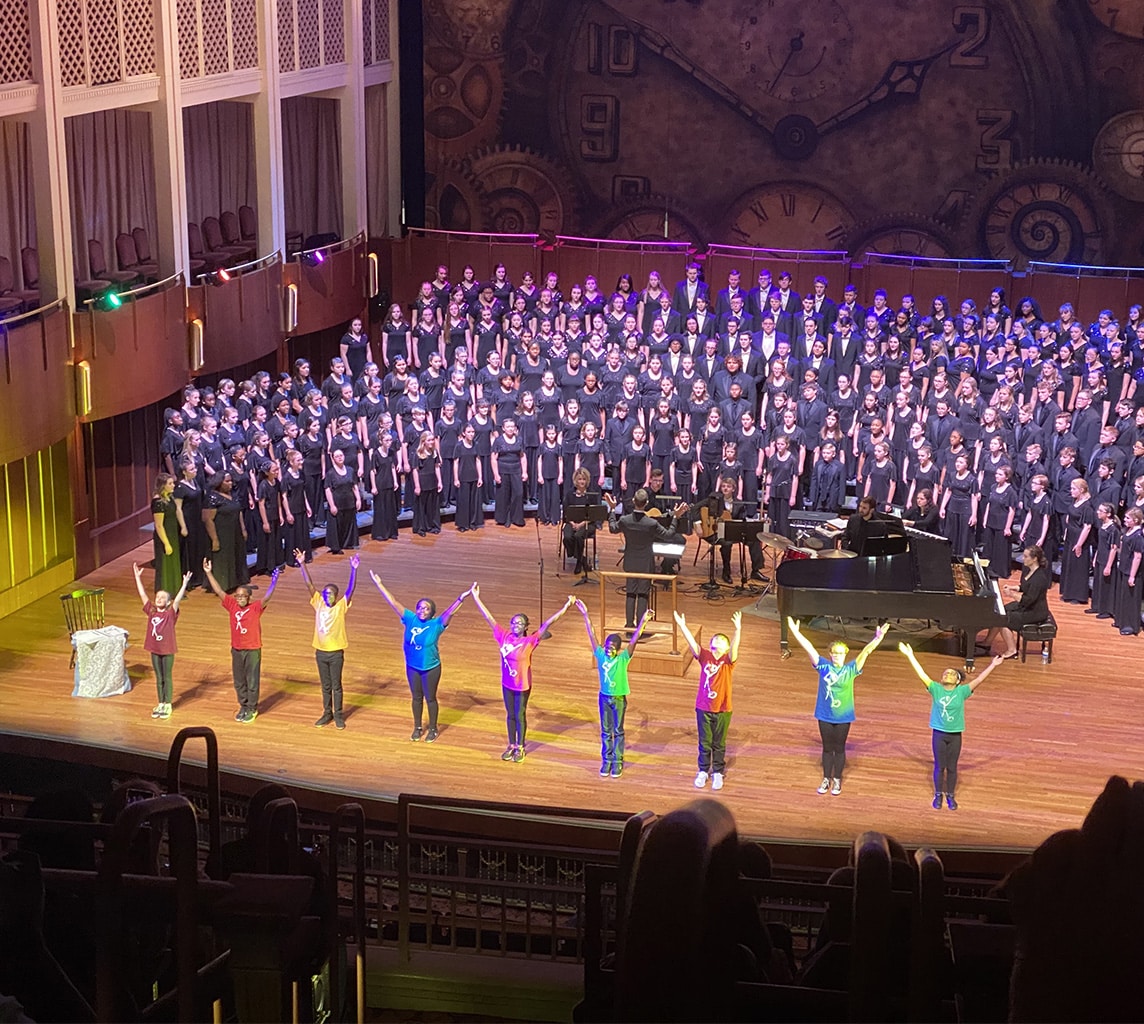 A large choir performs on stage with child dancers in colorful clothing holding hands in front. Detailed wall with large clock in the background.