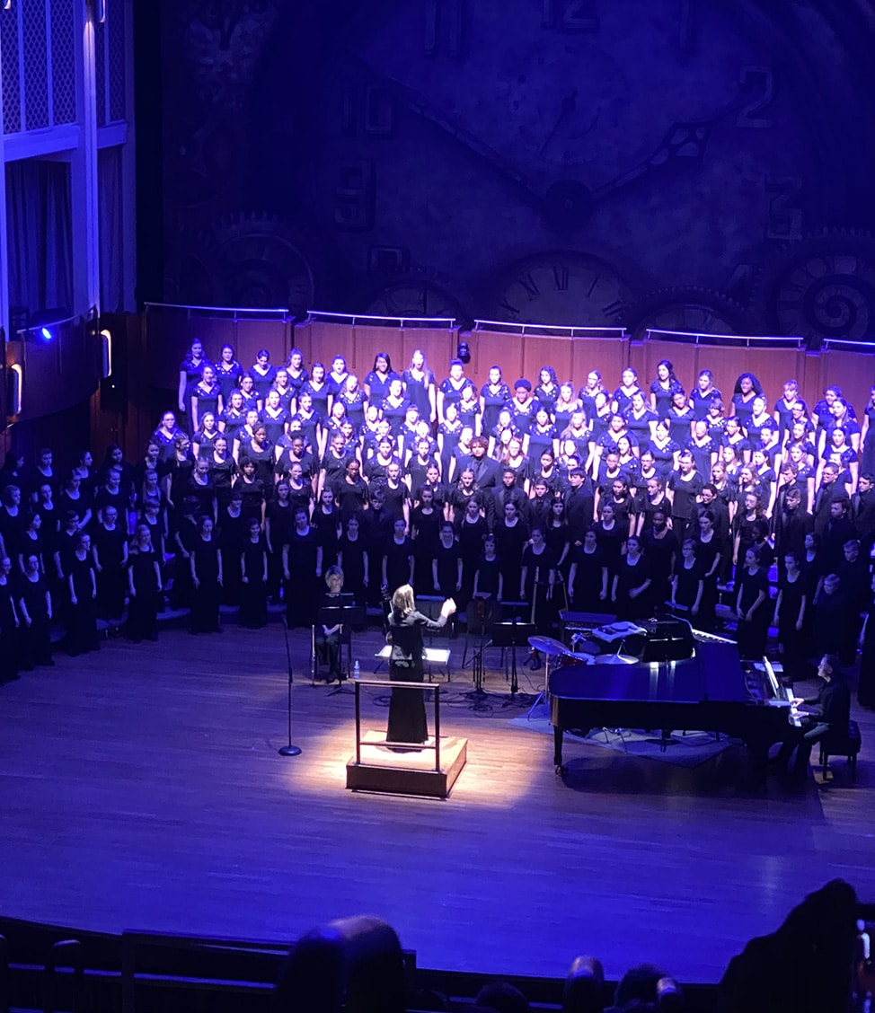 A conductor leads a large choir performing on stage. The choir members are dressed in black, and there is a piano on the right side. The backdrop features a large clock and mechanical gears.