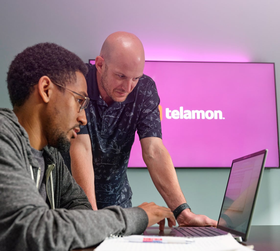 Two men in an office setting looking at a laptop screen, with a "telamon" logo displayed on a large screen in the background.