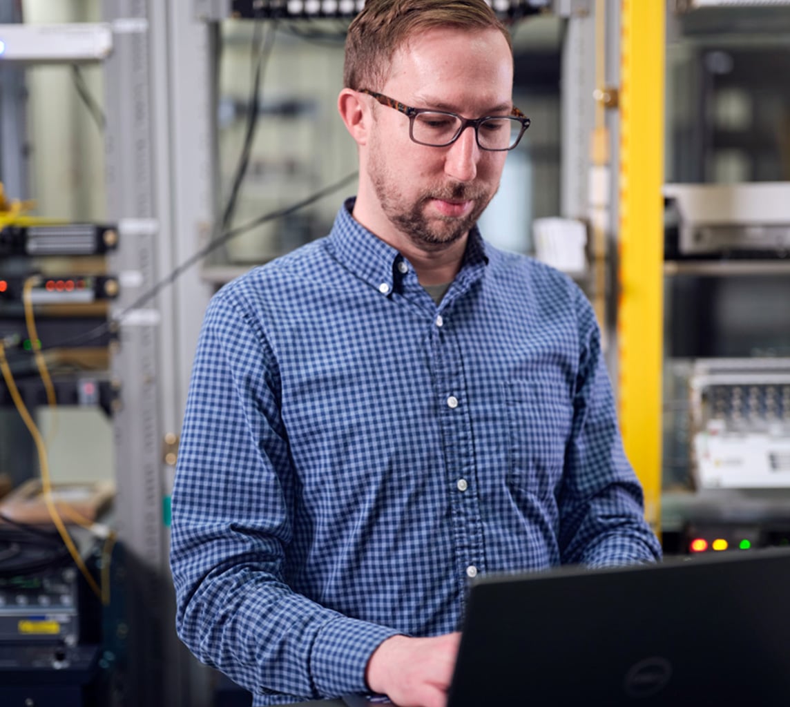 A person wearing glasses and a blue checkered shirt works on a laptop in a server room, surrounded by electronic equipment and wiring.