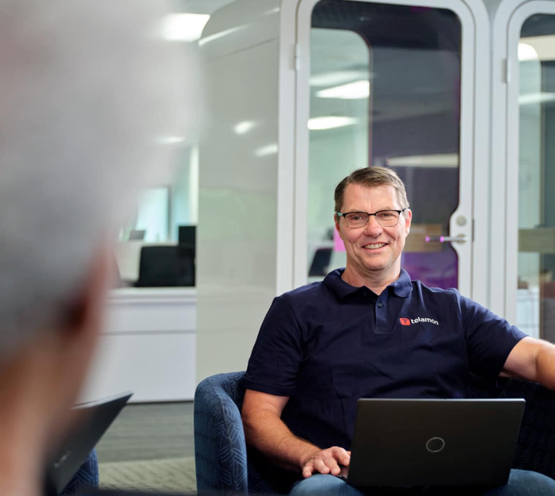 Person in a navy polo shirt sits in a modern office space, working on a laptop, and smiling at someone off-camera.