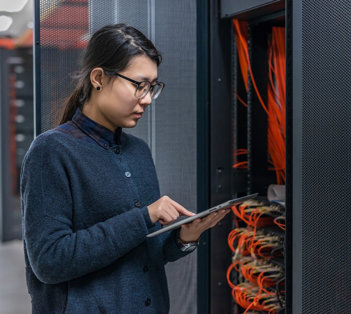 Person holding a tablet stands next to a server rack filled with cables and electronic equipment in a data center.