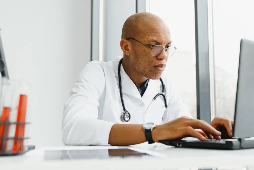 A person in a white lab coat and stethoscope types on a laptop while seated at a desk with lab equipment.