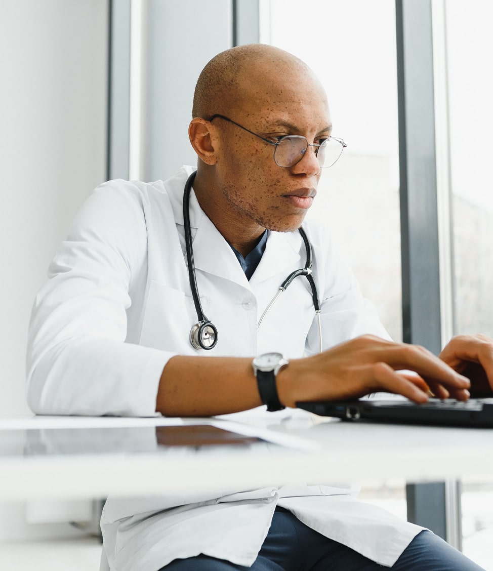 A doctor wearing glasses and a white coat with a stethoscope around his neck is typing on a keyboard at a desk.