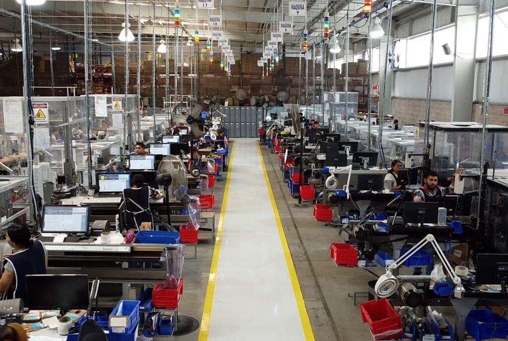 A busy factory floor with workers at individual stations, each equipped with computers and manufacturing equipment, under bright ceiling lights and a high central aisle.