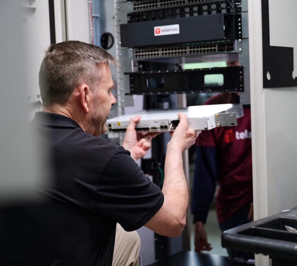 A technician is installing a hardware component into a server rack using both hands, while another person stands in the background.