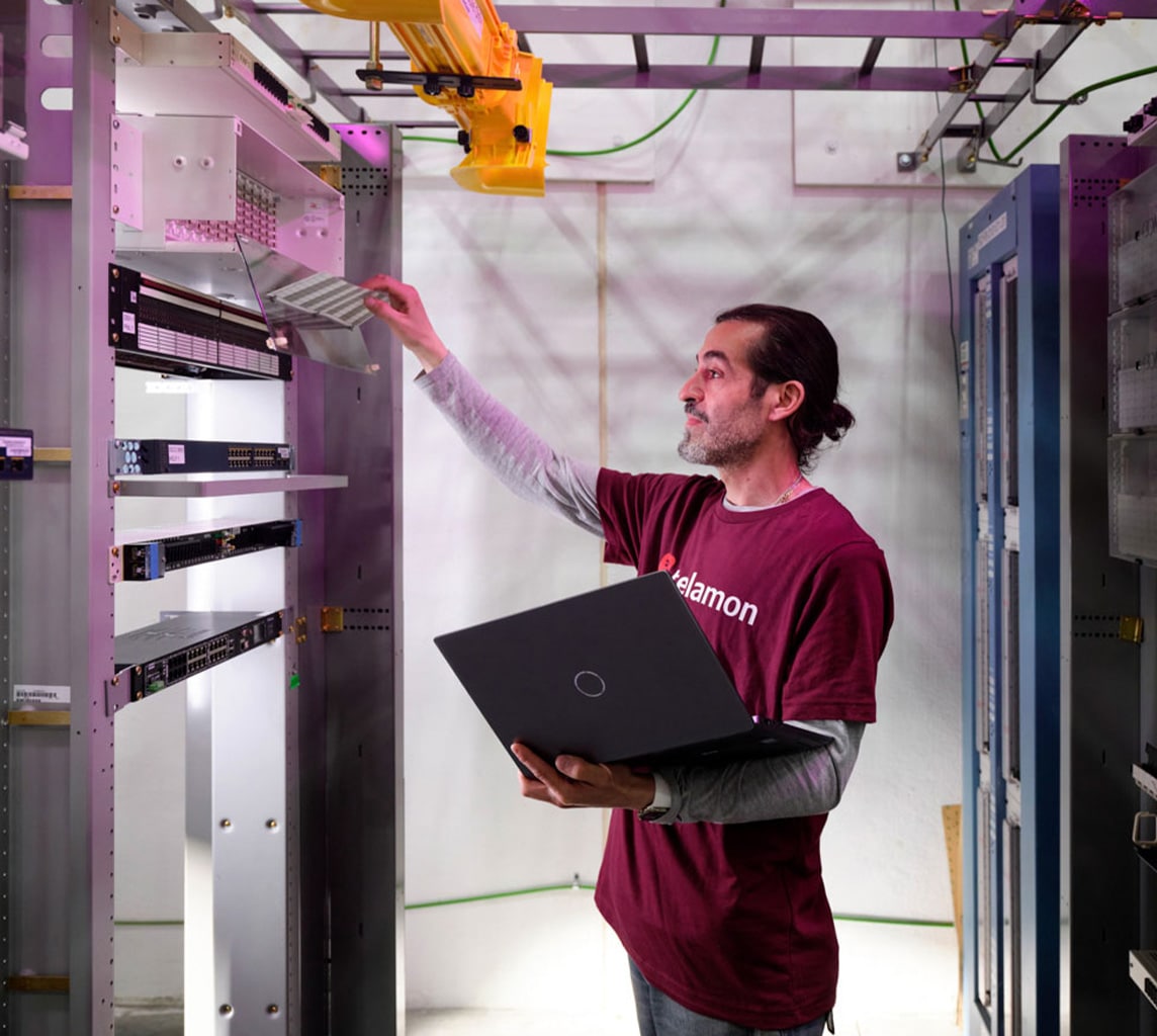 A person in a maroon shirt uses a laptop while working on a server rack in a server room.