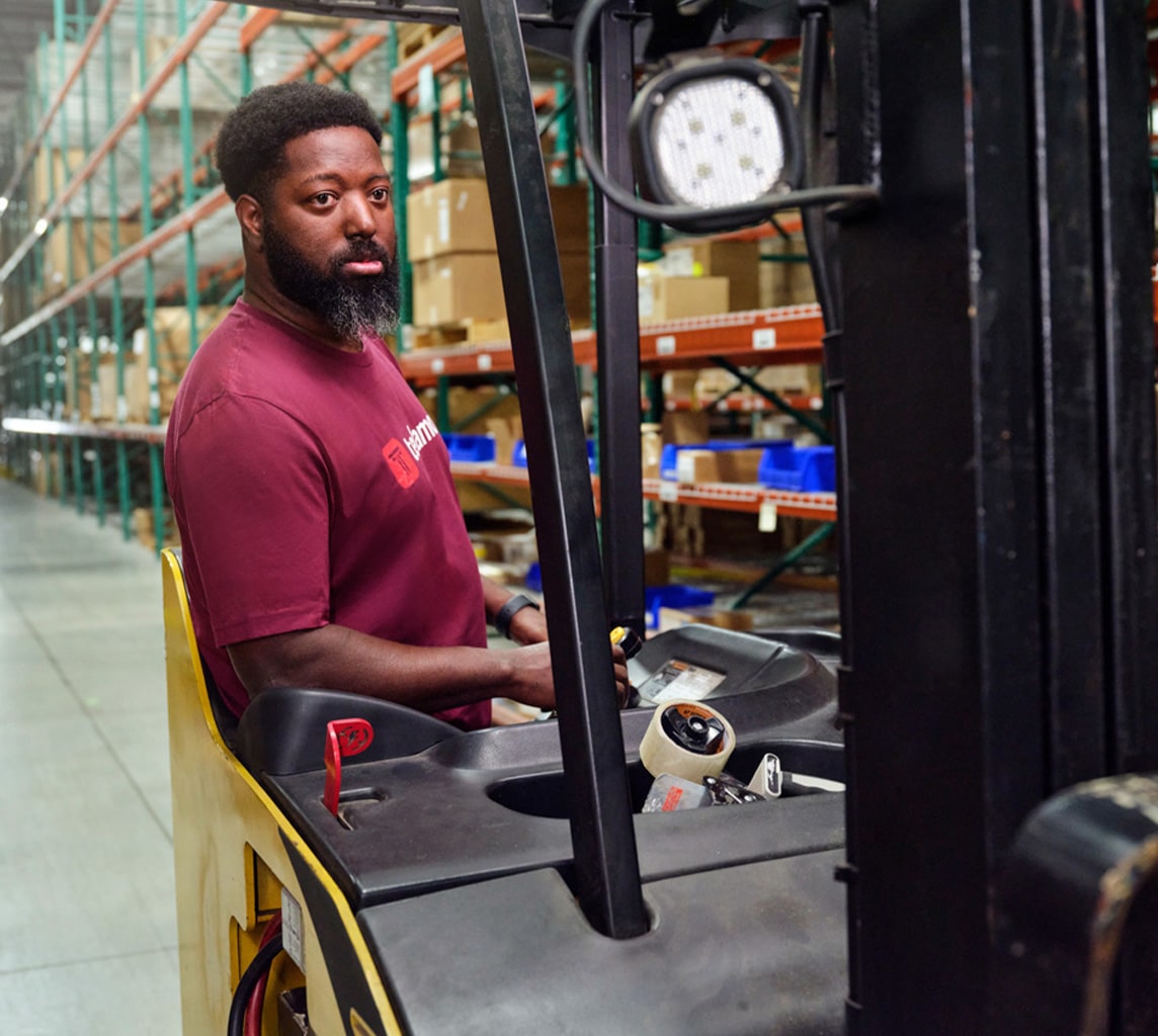 A man in a maroon shirt operates a forklift in a warehouse environment with shelves filled with boxes and items in the background.