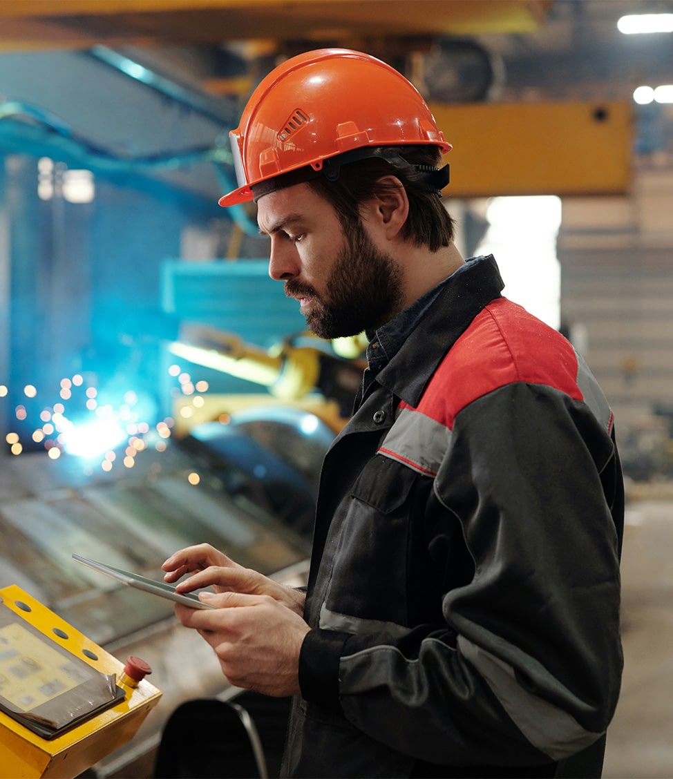 A male factory worker wearing a red hard hat and safety gear uses a tablet in an industrial setting with machinery and sparks in the background.