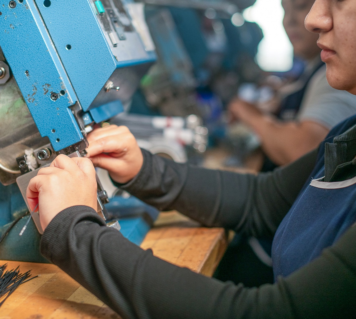 Close-up of a person's hands operating a blue industrial machine, focusing on inserting or adjusting components, with blurred workers and equipment in the background.