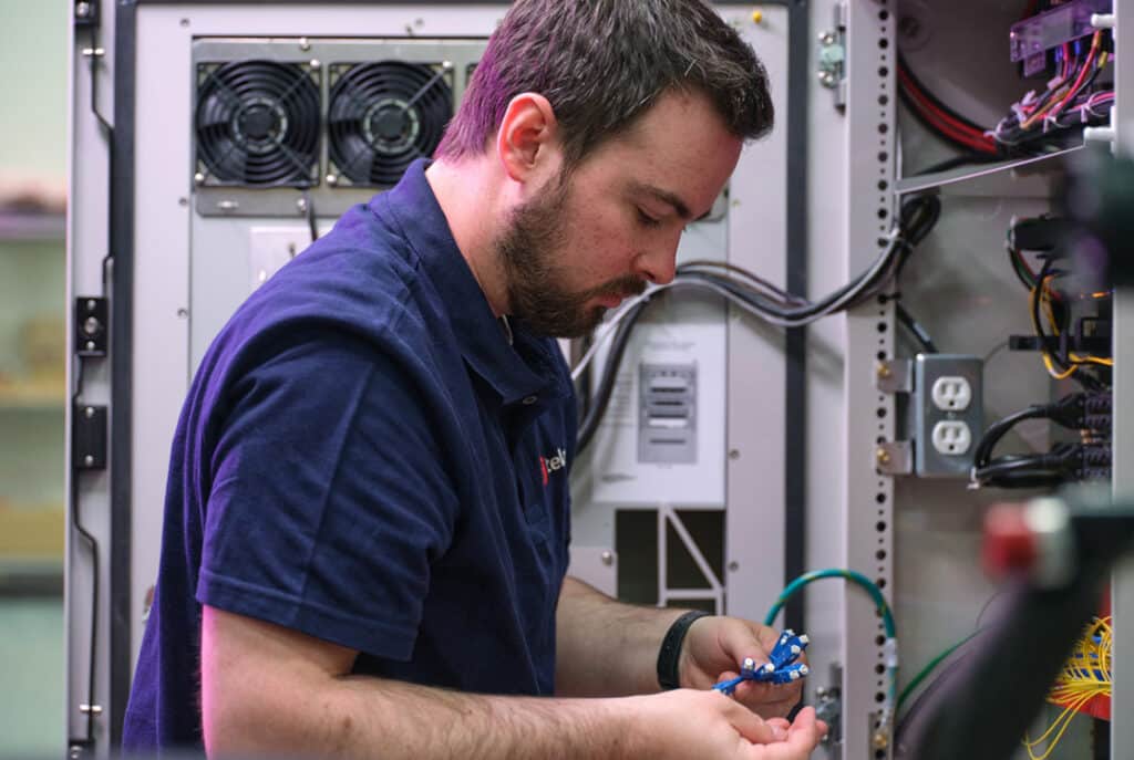 A man in a navy blue shirt works on wiring inside an electrical cabinet, surrounded by various cables and electronic components.