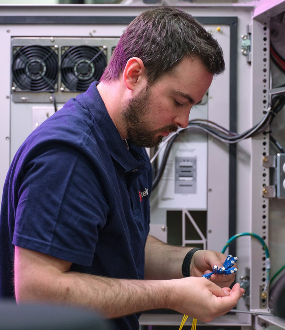 A man in a navy shirt is working on wiring within an open electrical panel, holding cables and tools in his hands.