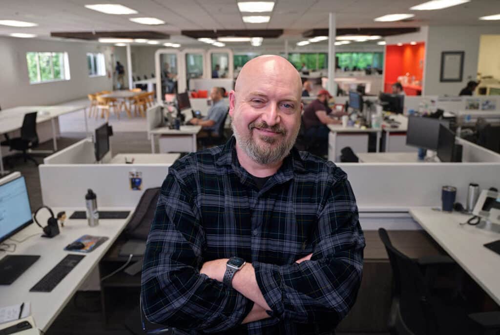 A bald man with a beard and wearing a plaid shirt stands with arms crossed in a modern open-plan office filled with workstations and employees.