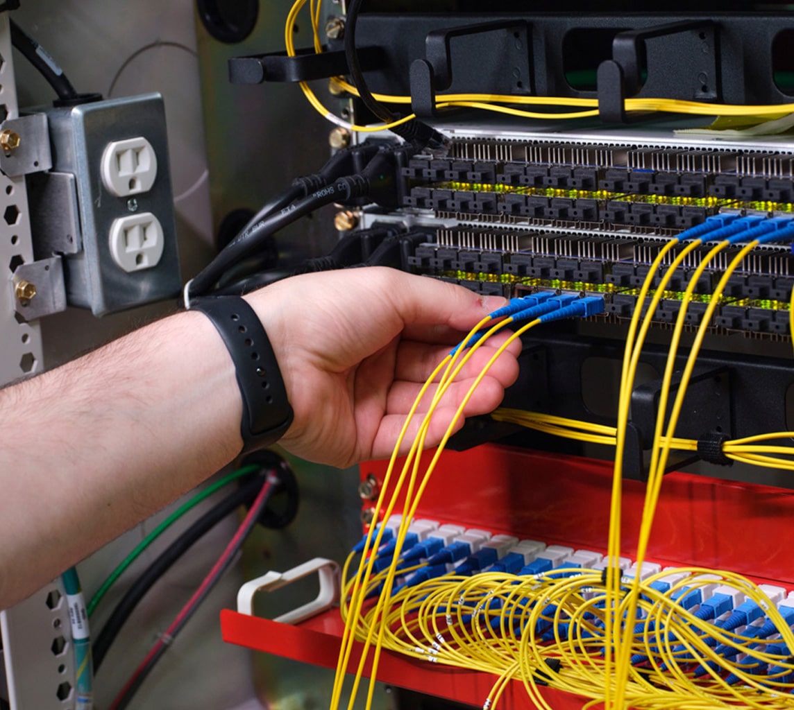 A hand plugs a yellow fiber optic cable into a patch panel, surrounded by an organized array of similar cables and electrical components.