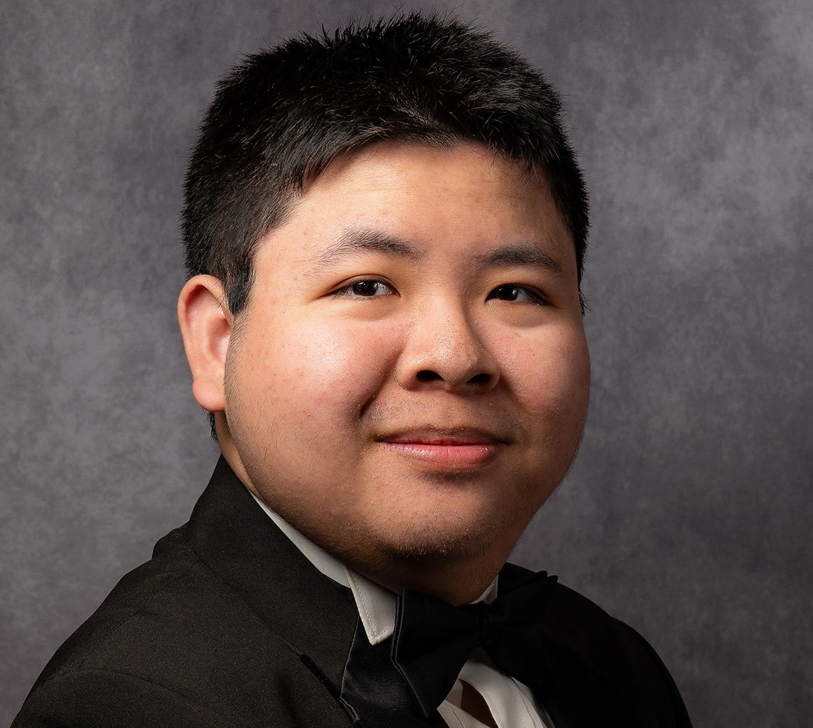 A person with short, dark hair wearing a black tuxedo and bow tie, posing against a gray textured background.