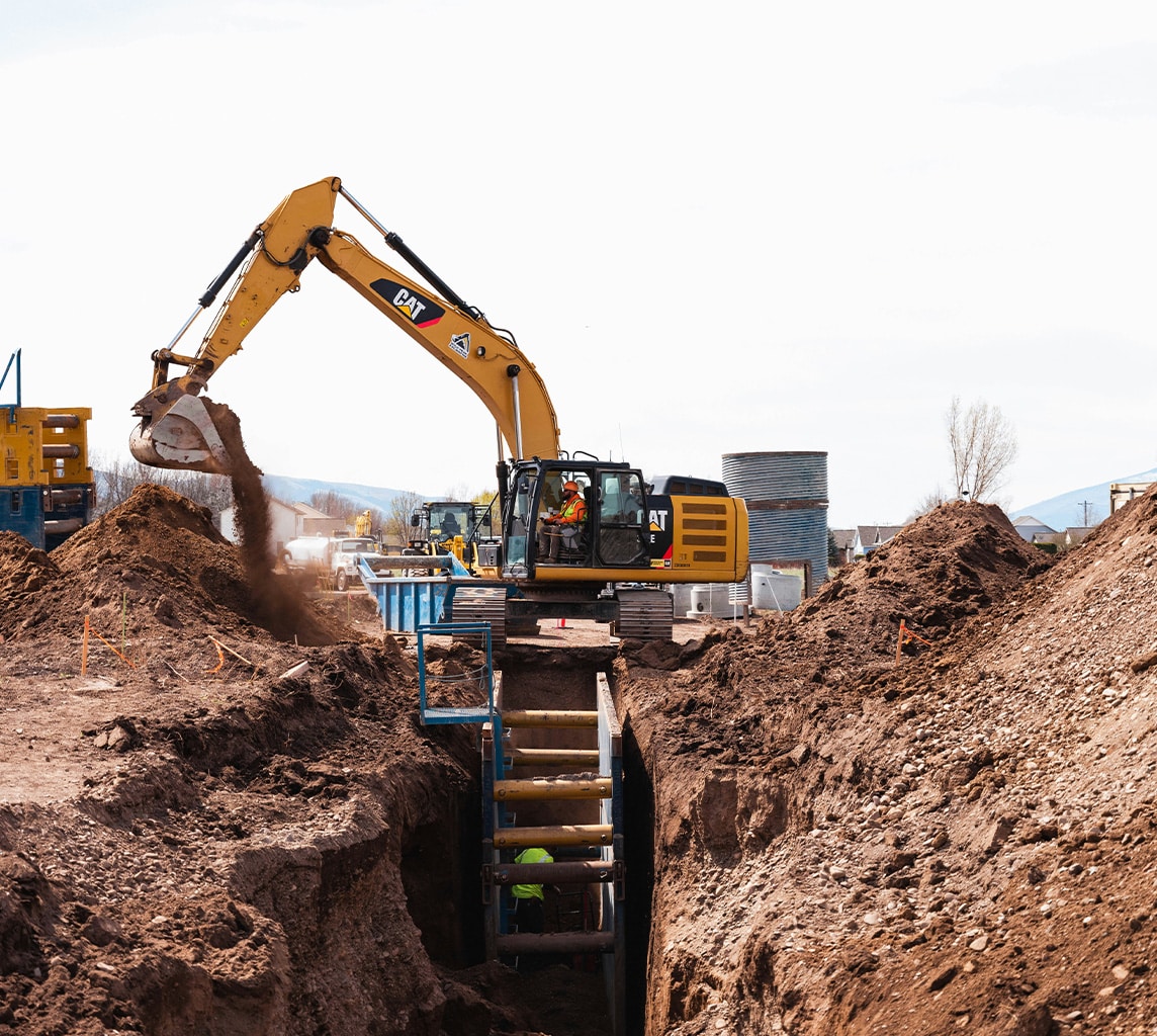 A construction site with an excavator digging a trench. Workers are visible inside the trench, and various construction materials and equipment are in the background.