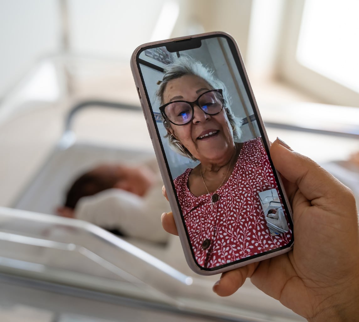 A person holds a phone showing a video call with an older woman. In the background, a newborn baby lies in a hospital bassinet.