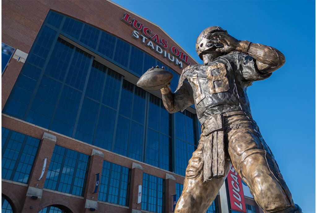 A bronze statue of a football player stands in front of Lucas Oil Stadium with the building's name visible on the facade.