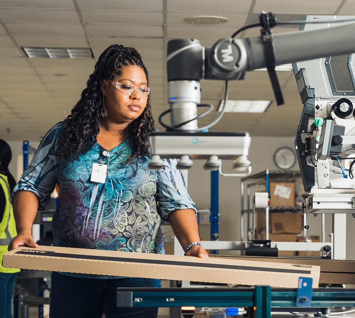 A person wearing safety glasses operates machinery in an industrial setting, holding a cardboard piece with a robotic arm in the background.