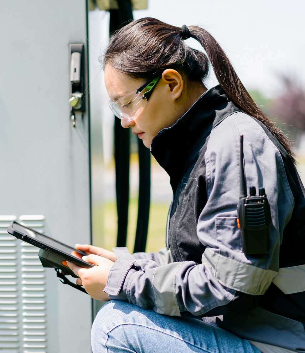 A woman wearing safety goggles and a jacket uses a tablet while kneeling beside equipment outdoors. A two-way radio is attached to her jacket.