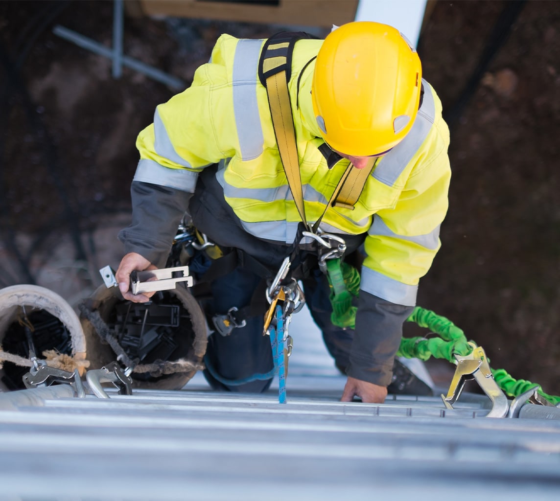 Worker in high-visibility jacket and yellow helmet climbing a ladder, carrying supplies with safety harness attached.