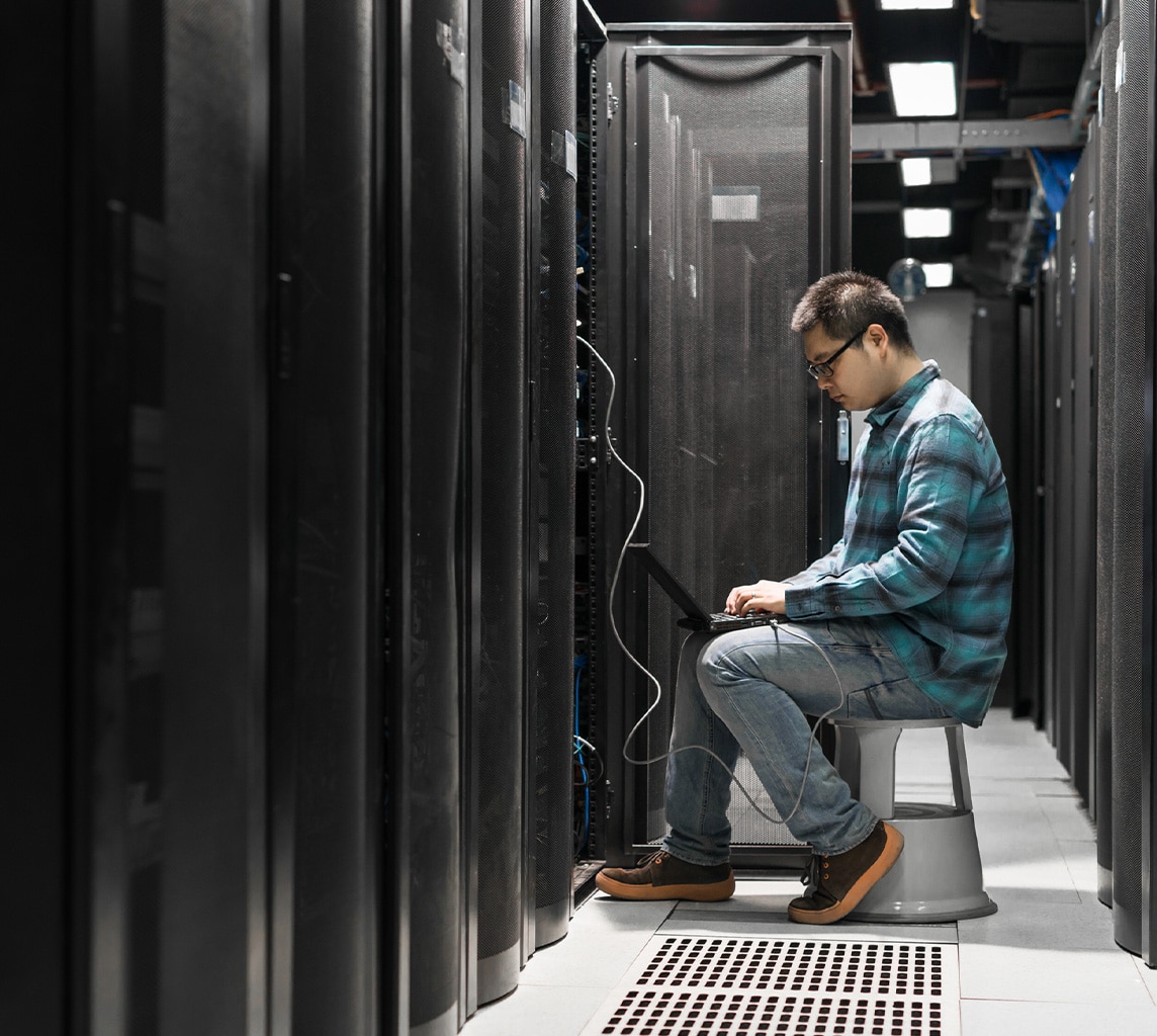 Person wearing glasses and a blue flannel shirt works on a laptop while sitting on a stool in a server room.