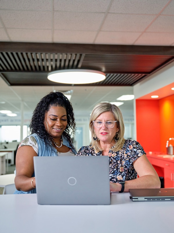 Two women collaborate on a project while looking at a laptop screen in a modern office setting. One woman has curly hair and wears a blue top; the other has blonde hair and wears glasses and a floral top.