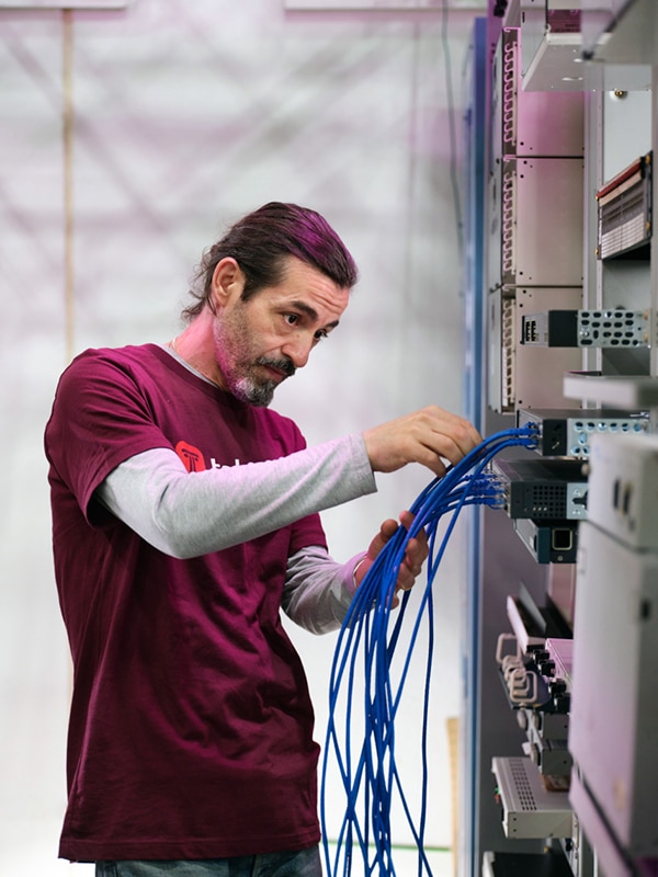 A person with a ponytail, wearing a maroon t-shirt over a grey long-sleeve shirt, is plugging blue cables into server equipment in a data center.