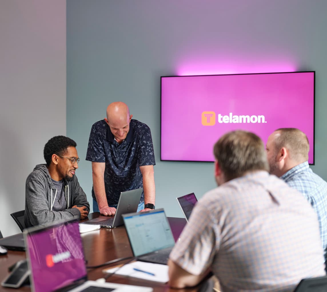 Four men are having a discussion around a conference table, with laptops open. A screen behind them displays the logo and name "Telamon.