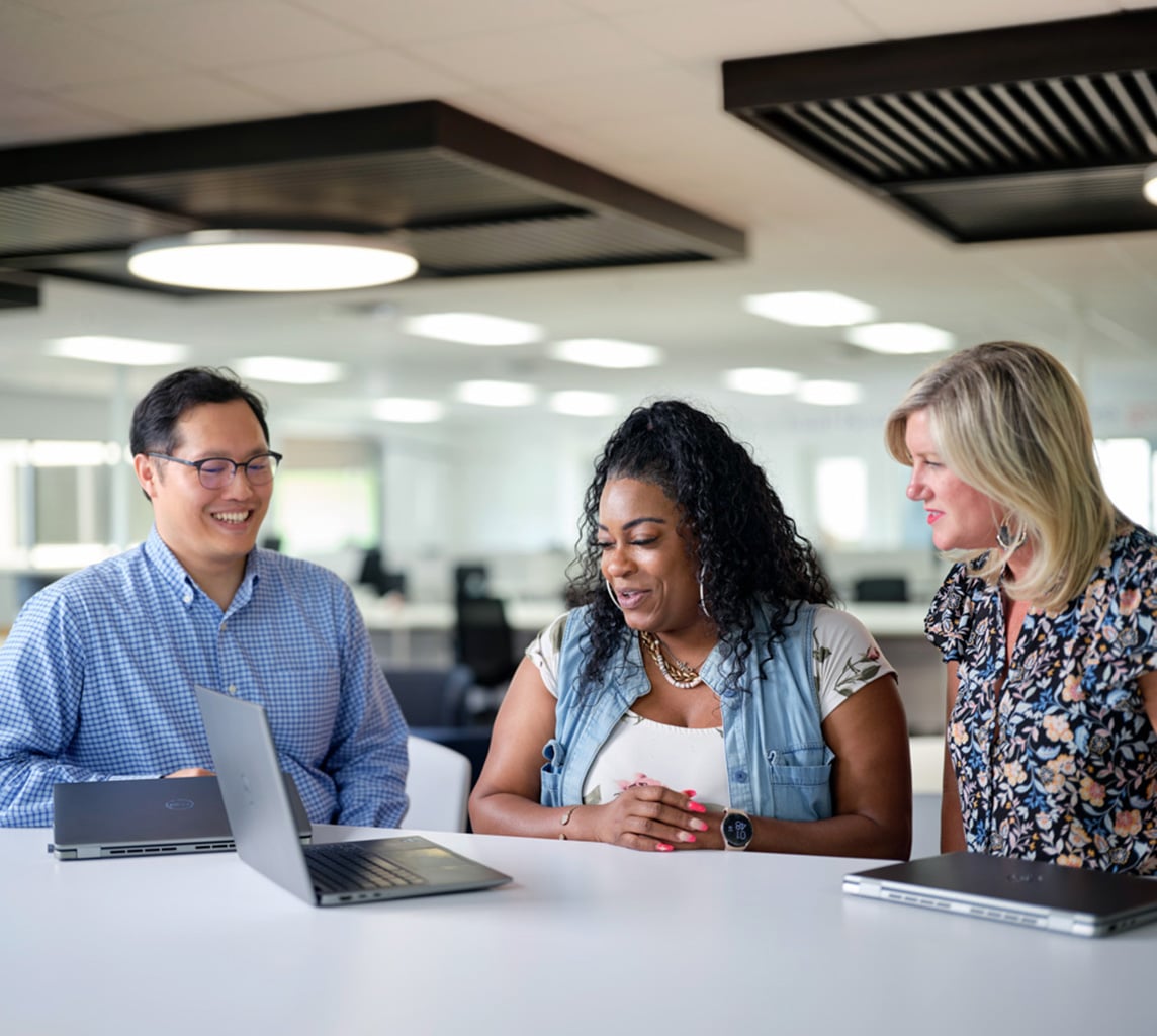 Three people are gathered around a table in an office, engaging in discussion with open laptops in front of them.