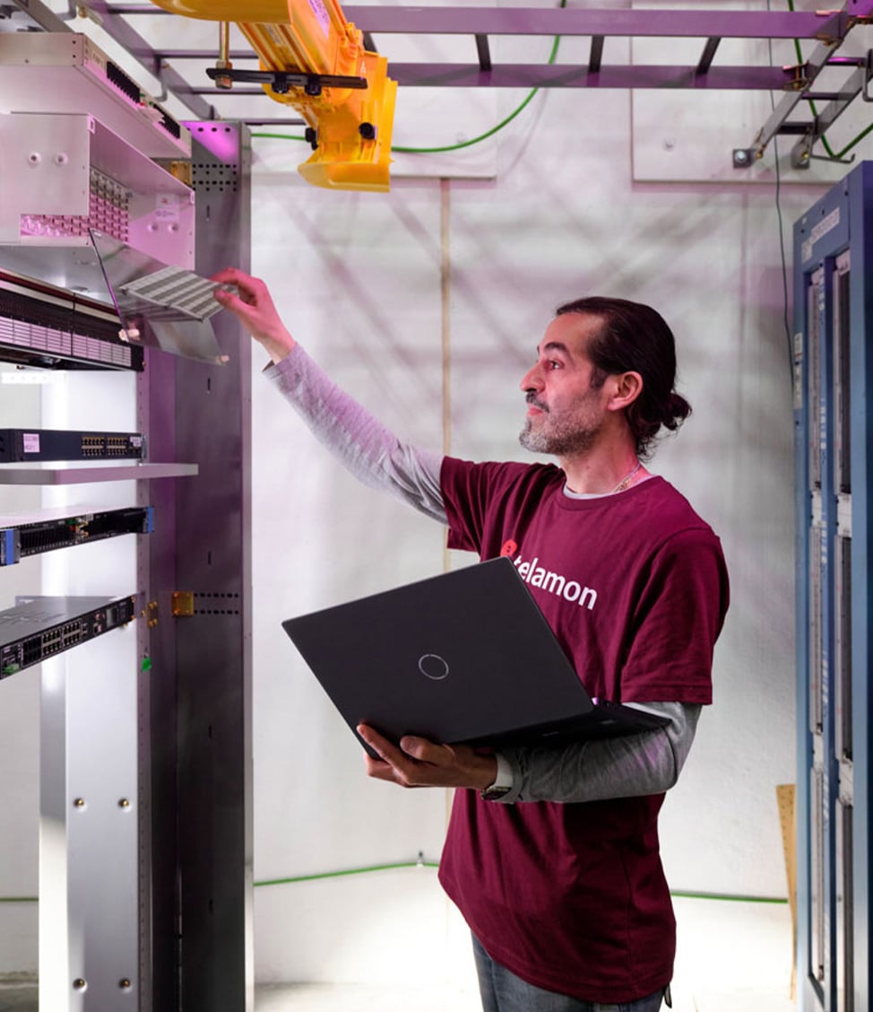 A person in a maroon shirt inspects server equipment in a data center while holding a laptop.