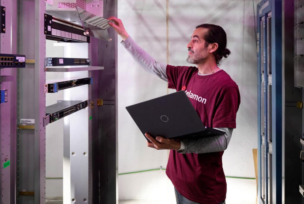 An individual in a maroon shirt is working in a server room, holding a laptop in one hand and adjusting a server rack with the other hand.