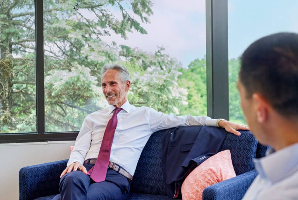 Two men in business attire are sitting and talking on a blue couch near a large window with a scenic outdoor view. One man, with gray hair and a red tie, is smiling while the other listens attentively.