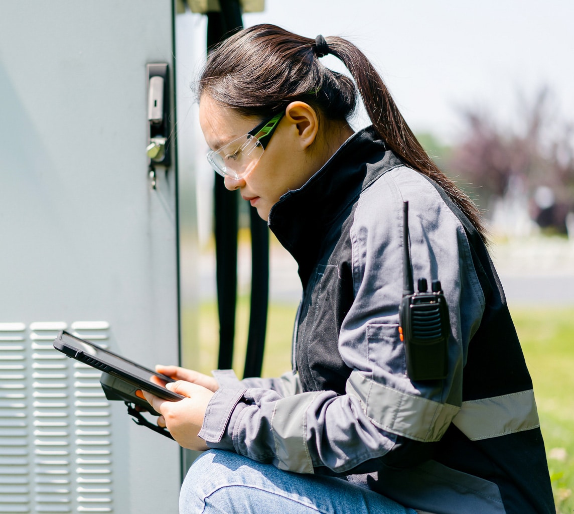 A woman technician wearing protective glasses and a two-way radio on her sleeve, is working outside with a tablet device near an electrical cabinet.