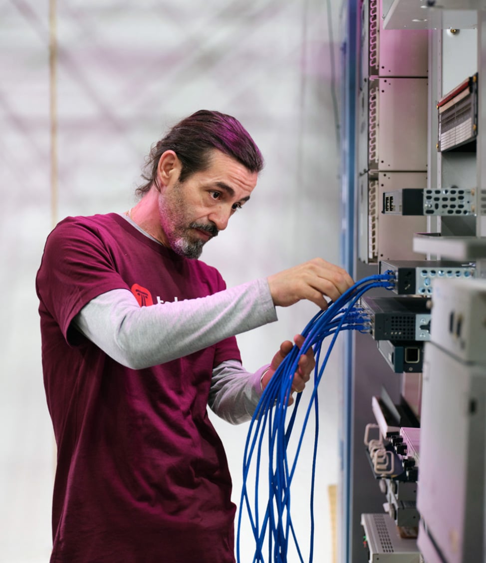 A man with long hair and a beard, wearing a maroon shirt, is organizing blue network cables connected to a server rack.