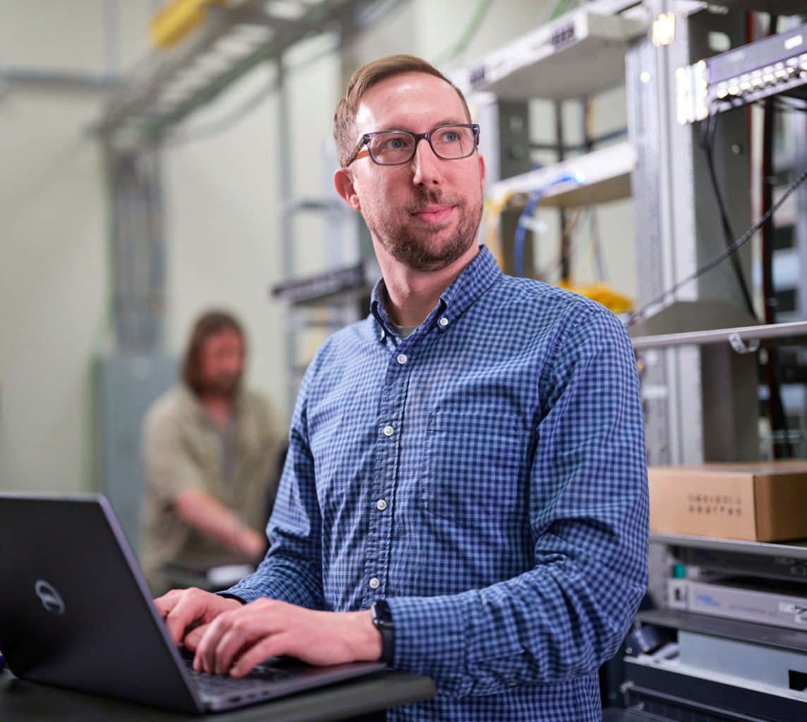 A man in glasses and a blue checkered shirt works on a laptop in a server room, with another person working in the background.