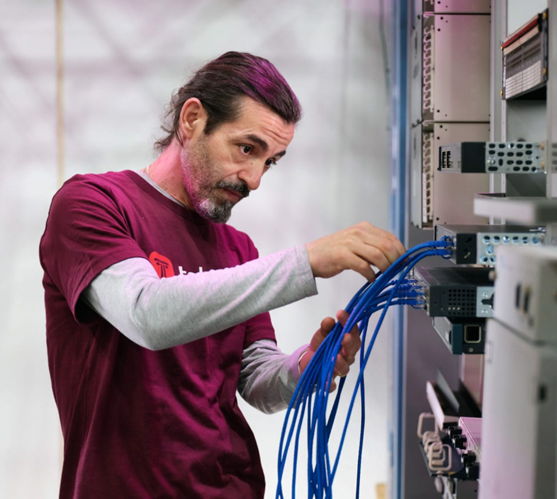 A man with long hair and a beard works with blue network cables in a server room.