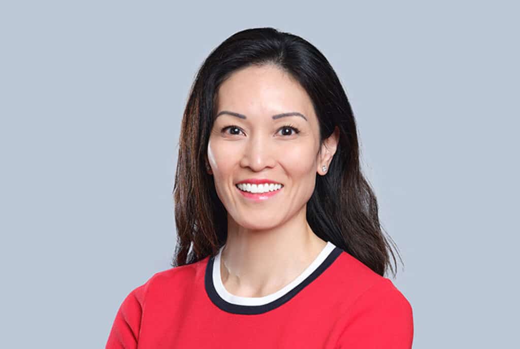 A woman with long dark hair wearing a red top smiles, standing against a plain blue background.