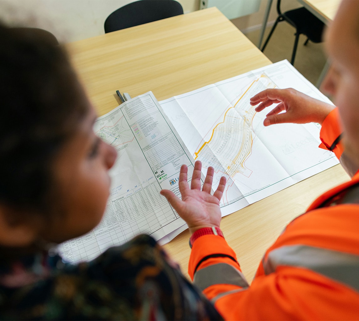 Two individuals are discussing a blueprint or map spread out on a table. One is pointing at a specific section on the document. Both are wearing work attires.