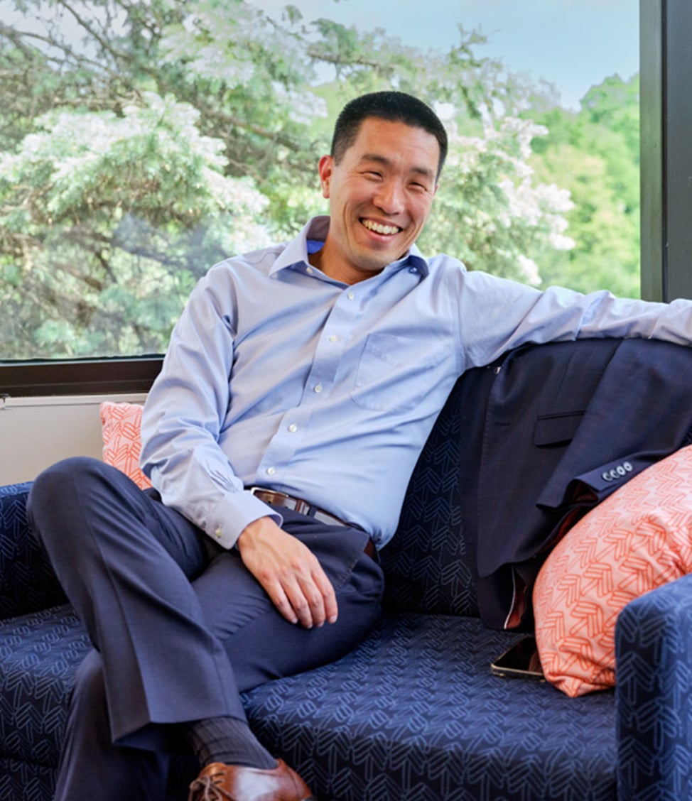 A man in a blue shirt and dark pants sits on a couch, smiling with a suit jacket draped over the backrest and trees visible through a window behind him.